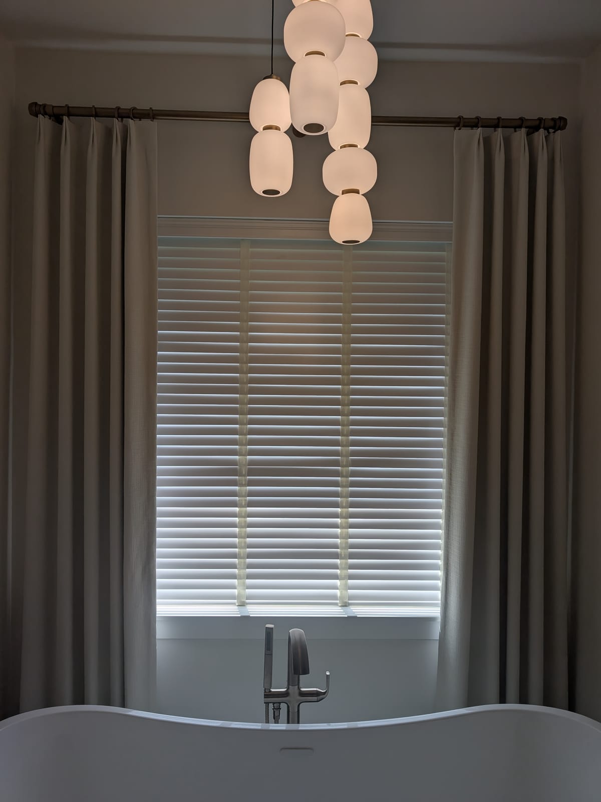 White two-inch blinds framed by taupe drapery panels above a freestanding tub with pendant lights in a modern bathroom