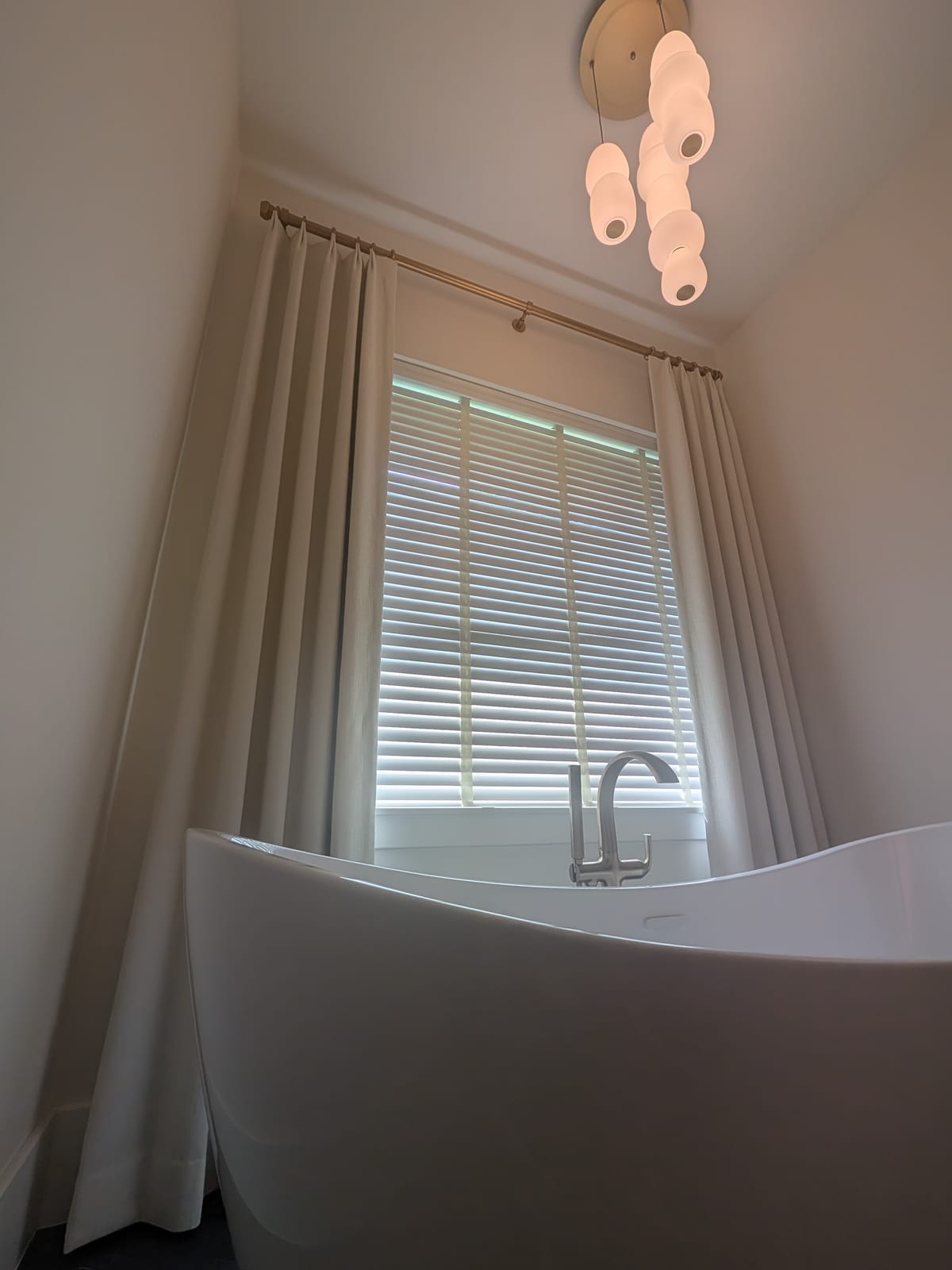 Freestanding soaking tub beneath a window with white blinds and floor-length taupe draperies