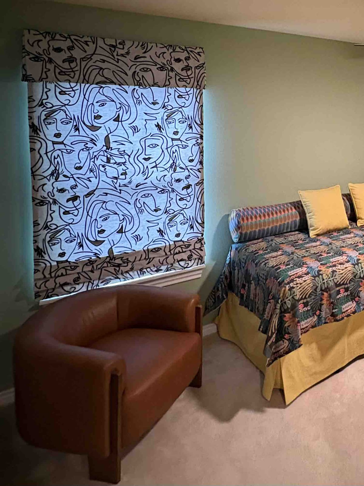 Custom roman shade with matching valance in a bold line-art faces fabric beside a leather chair in a sage green guest bedroom